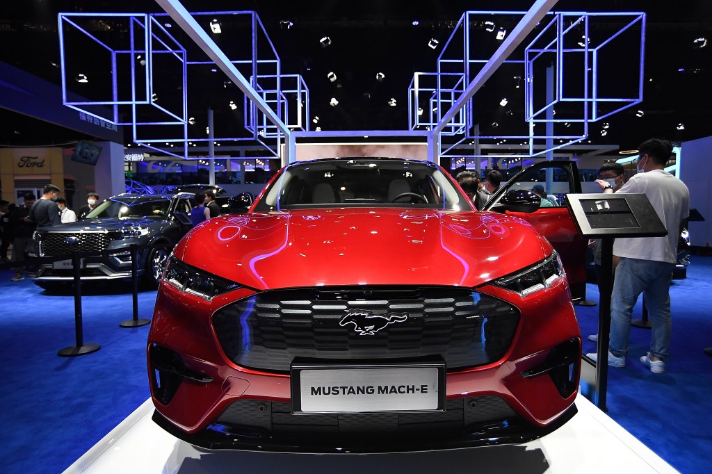 A red 2021 Ford Mustang Mach-E sits on the floor of a car show. The Mach-E is one of Ford's new electric vehicles. 