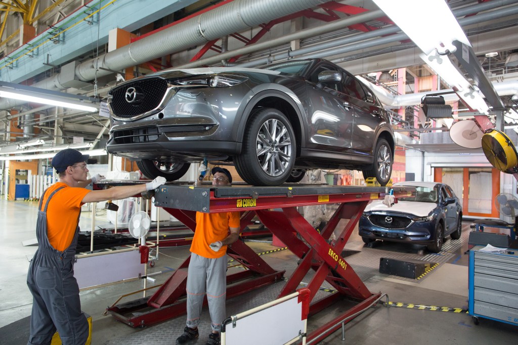 Workers affix parts to the underside of a gray Mazda CX-5 sports utility vehicle (SUV) on the assembly line at the Mazda Sollers Manufacturing Rus LLC plant