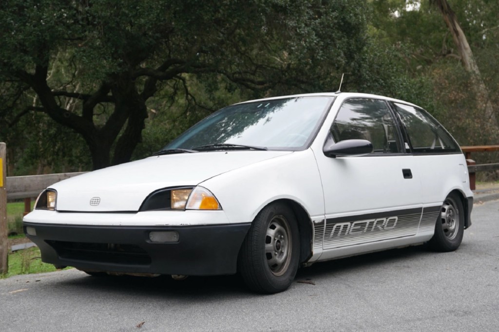 A modified black-and-white 1990 Geo Metro parked by a forest