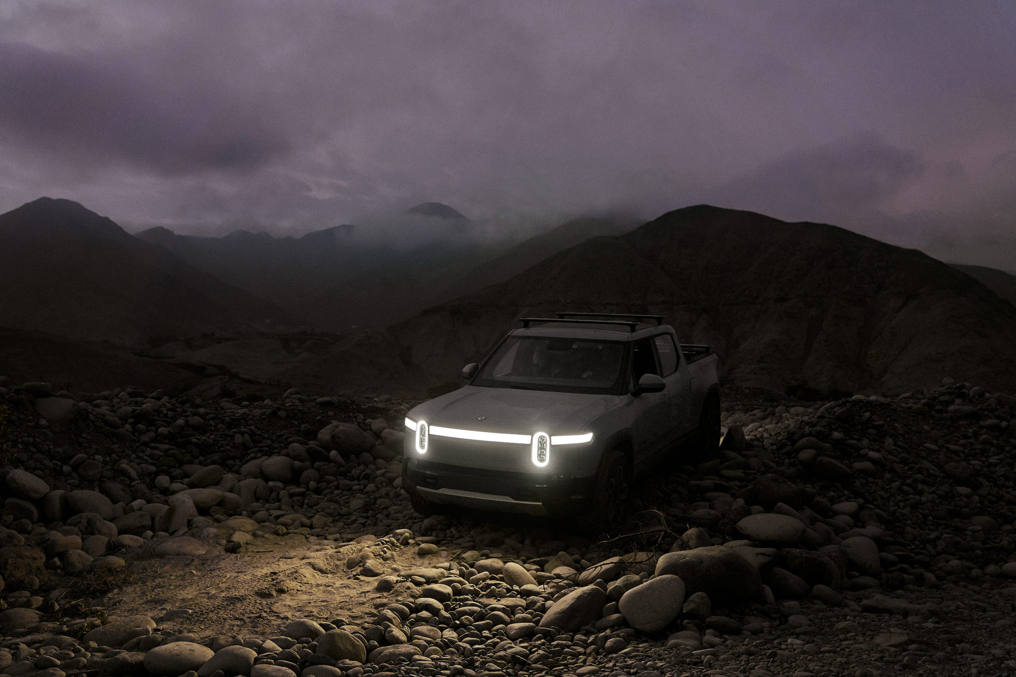 A gray Rivian R1T parked on rocks in front of cloud-shrouded mountains at dusk