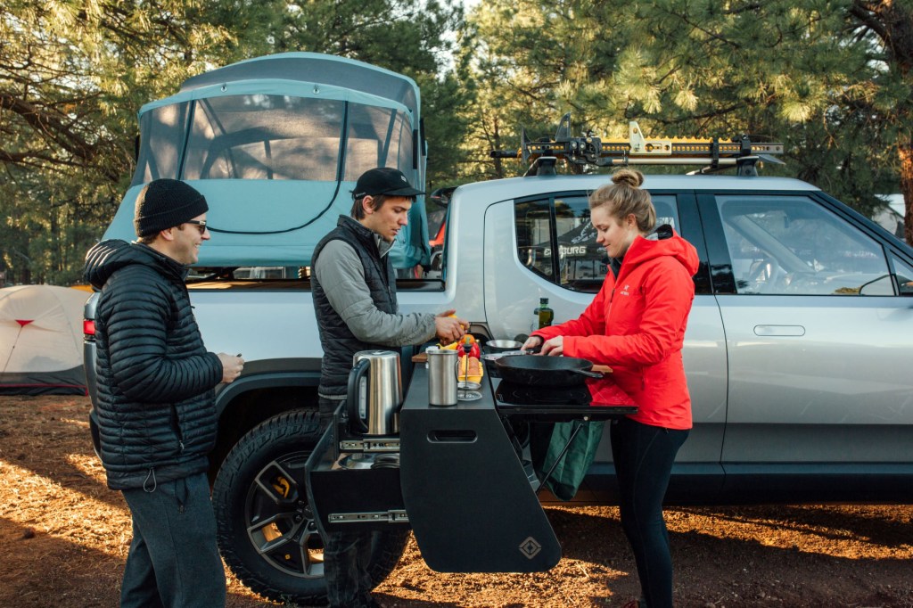 Campers next to a silver Rivian R1T with a bed-mounted tent and a slide-out camp kitchen