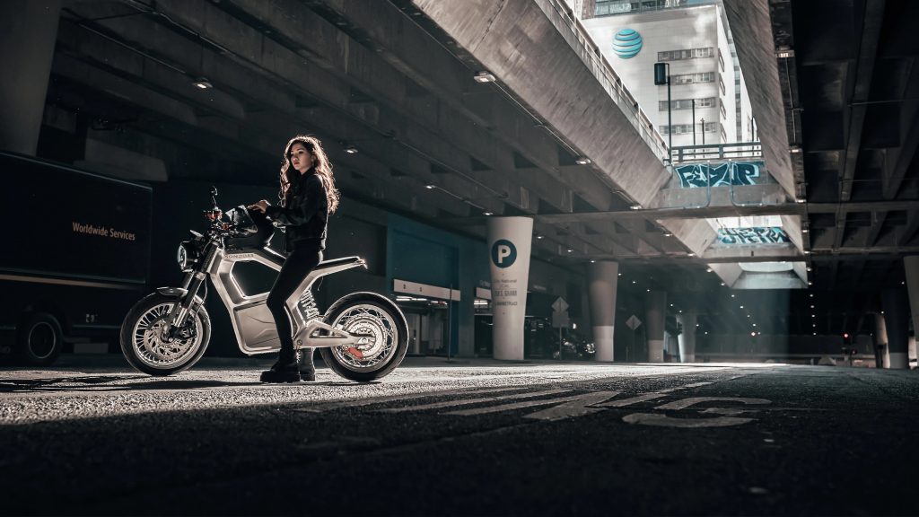 A black-clad rider on a silver Sondors Metacycle on a city street underneath a bridge