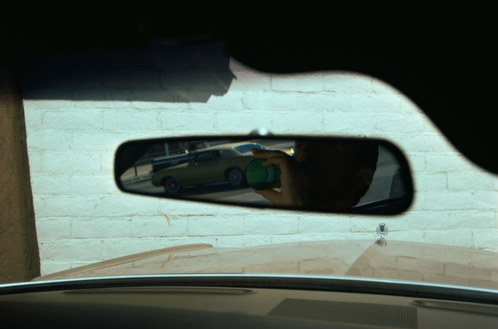 A reflection, in the rear-view mirror of a car, of a man drinking from a bottle.