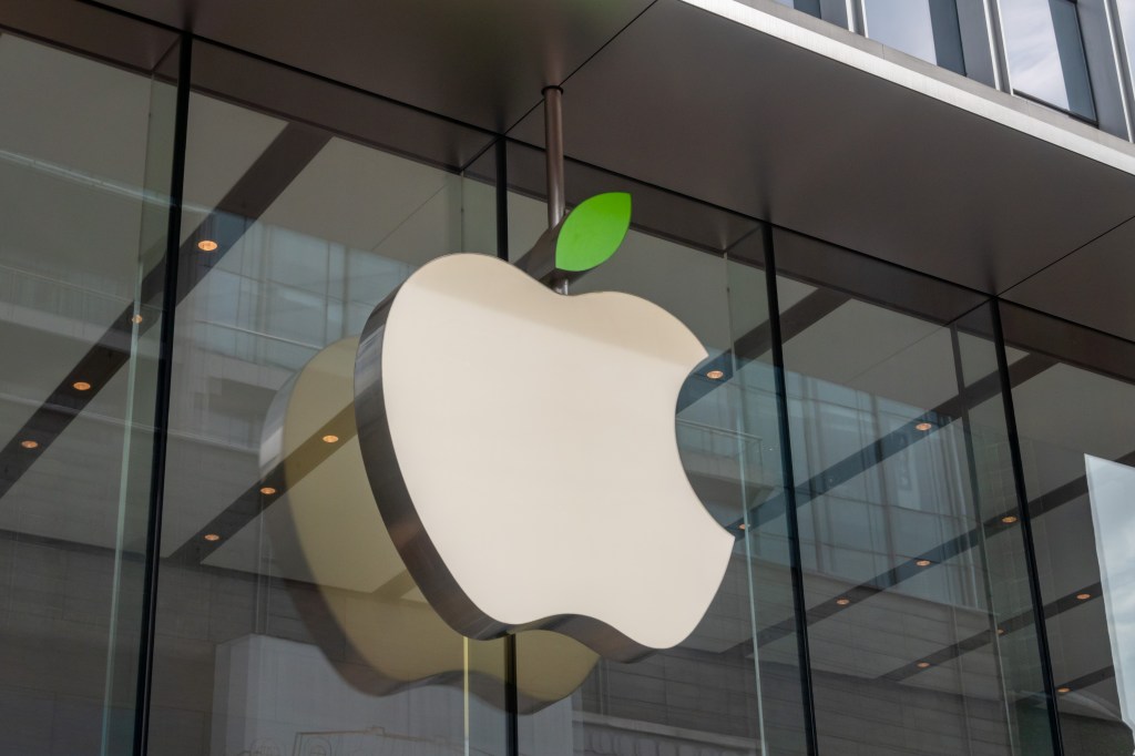 An Apple logo hanging from a glass wall outside one of their stores.