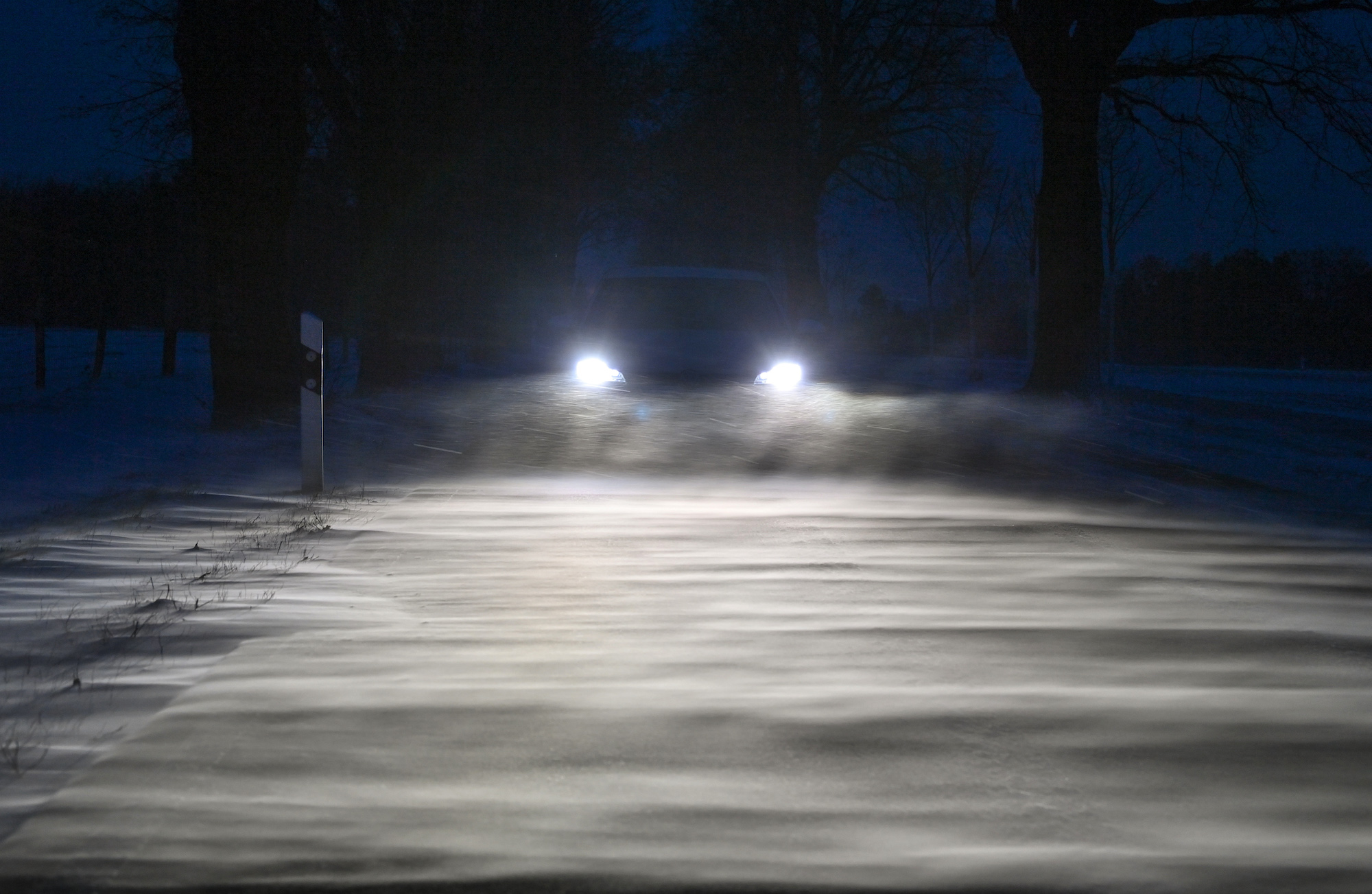 A car's headlights shine across a snow-covered road in the thick of evening snow