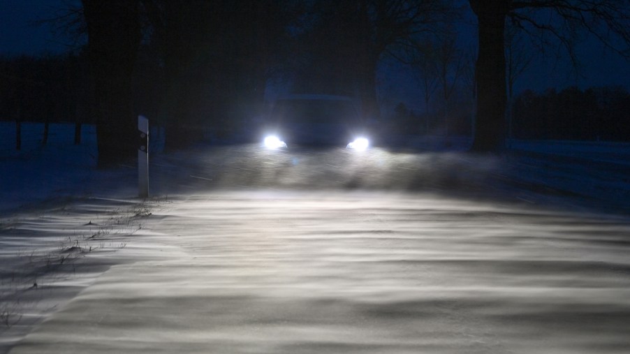A car's headlights shine across a snow-covered road in the thick of evening snow