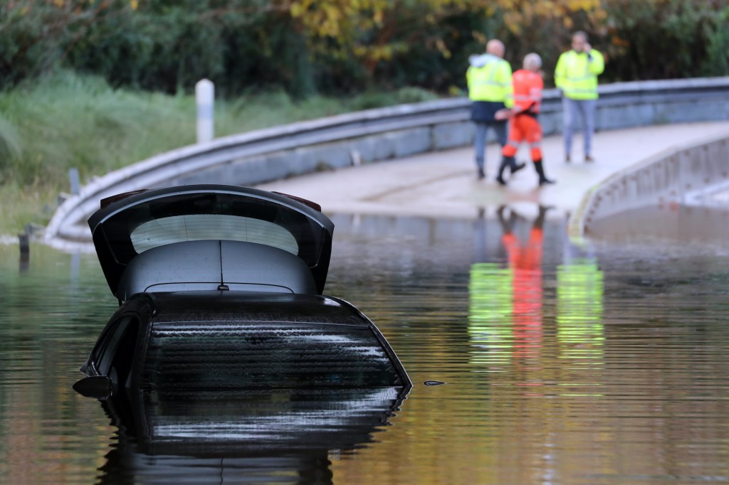Rescuers walk by a car partially submerged in the water on a flooded road.