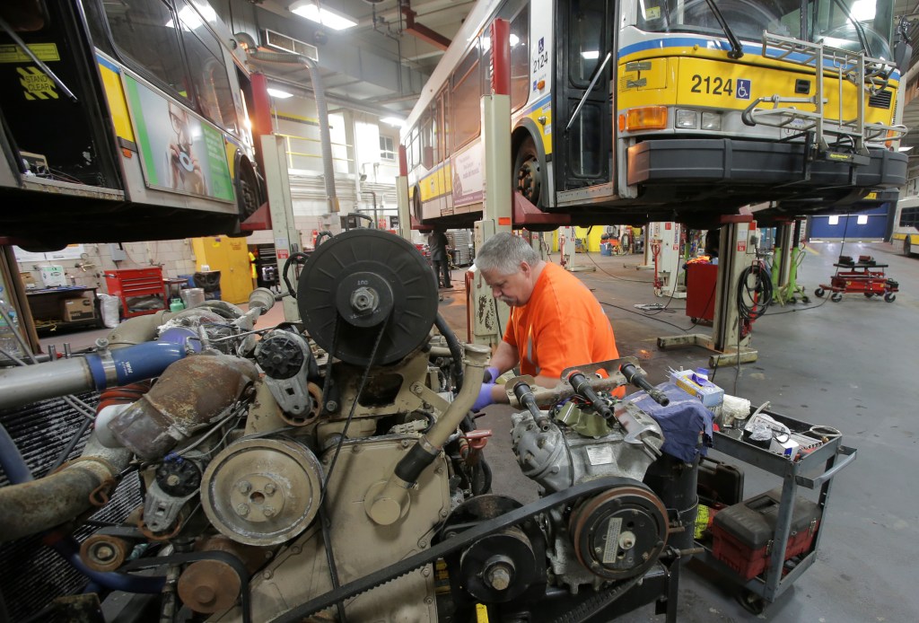 A machinist in an orange tee shirt and blue gloves rebuilds a large bus engine