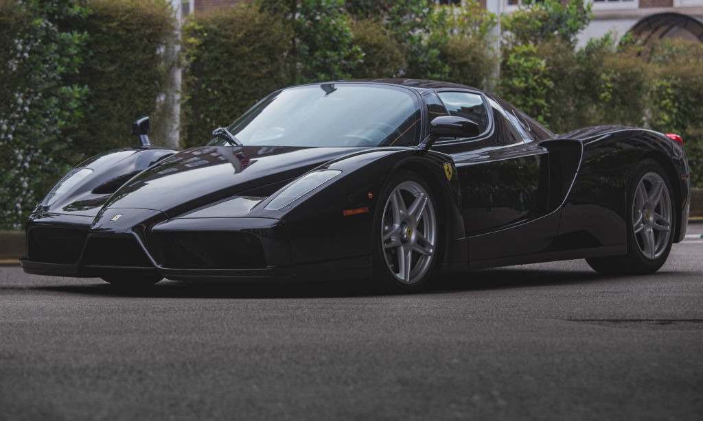 A black Ferrari Enzo sits in the shade against a row of green hedges