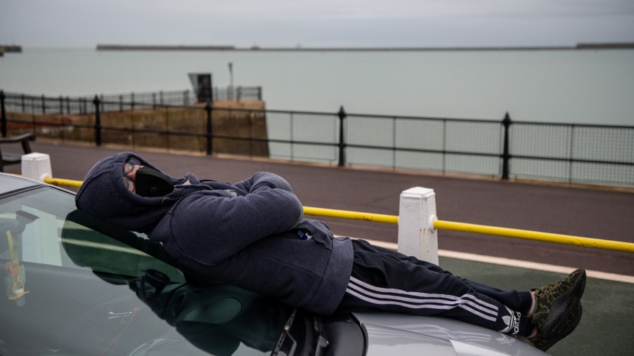 A man wearing a facemask sleeps on the hood of his car, overlooking a large body of water