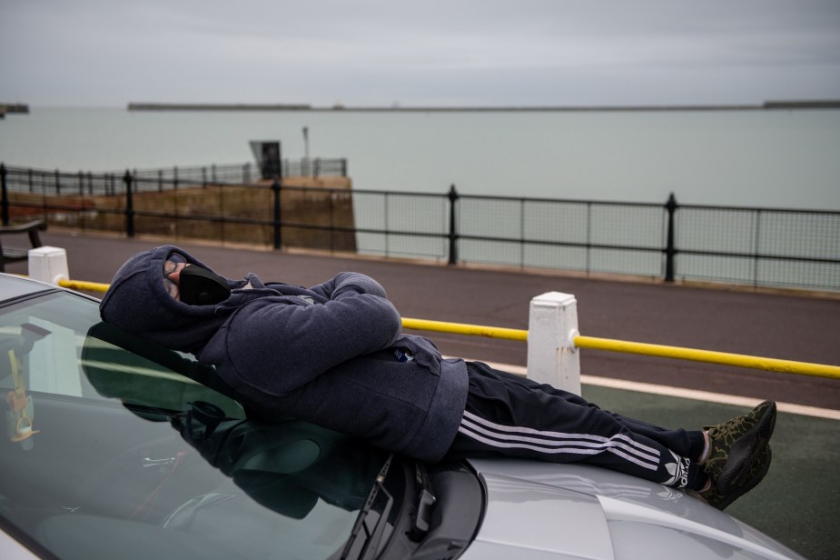 A man wearing a facemask sleeps on the hood of his car, overlooking a large body of water