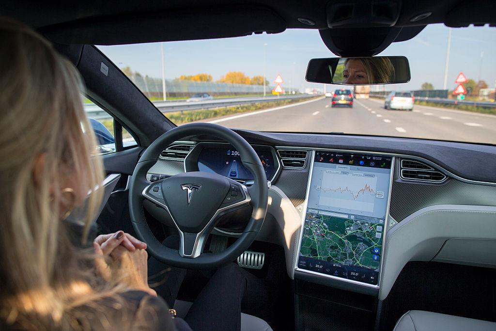 A woman sits in the driver seat of a Tesla, hands off the wheel, while autopilot guides the car down a road