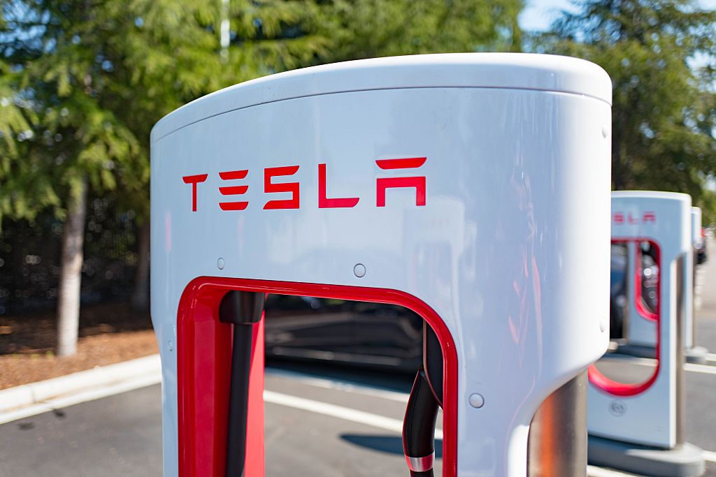A white Tesla EV supercharger with red lettering sits in the sun