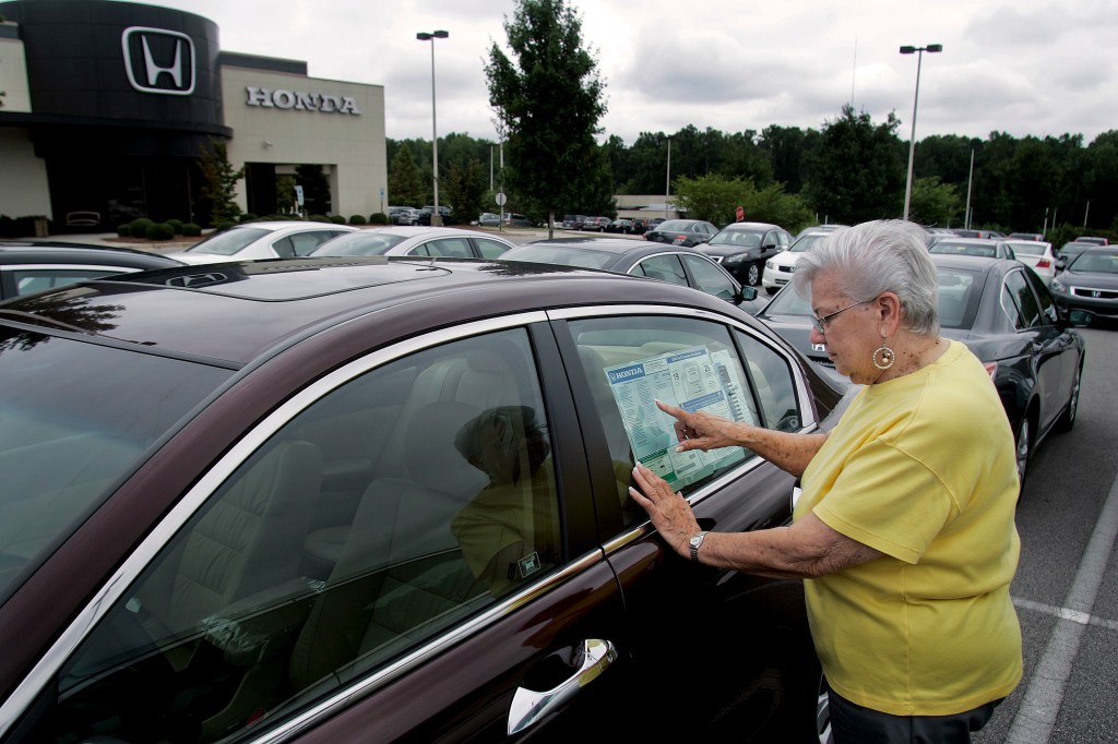 A woman with short silver hair in a yellow tee shirt examines the window sticker of a maron sedan on a sales lot