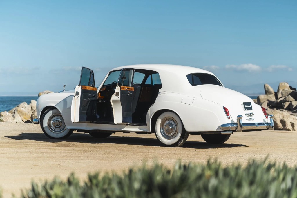 The rear 3/4 view of a white 1965 Rolls-Royce Silver Cloud III with its doors open on a beach