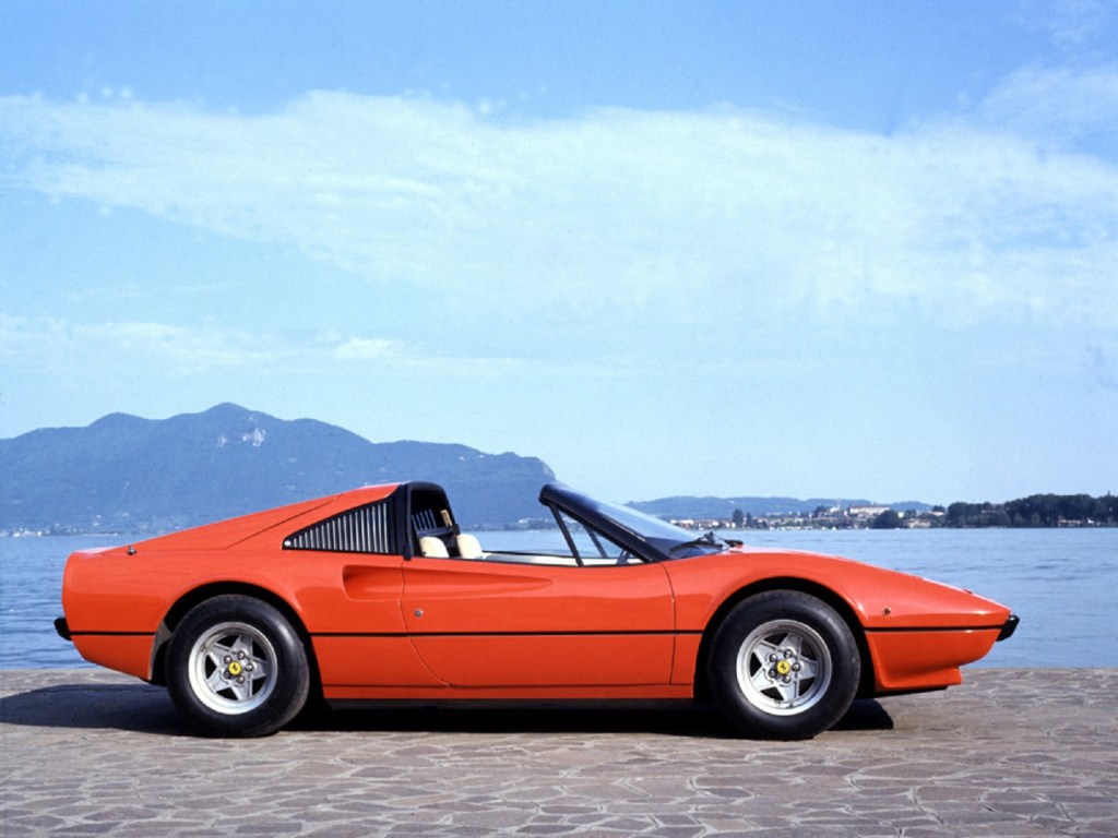 The side view of a red 1977 Ferrari 308 GTS with its roof off parked next to a lake