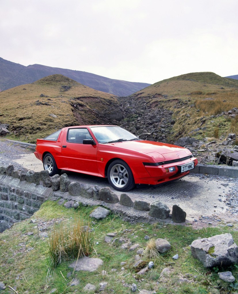 A red 1987 Mitsubishi Starion on a stone bridge among rolling mossy hills