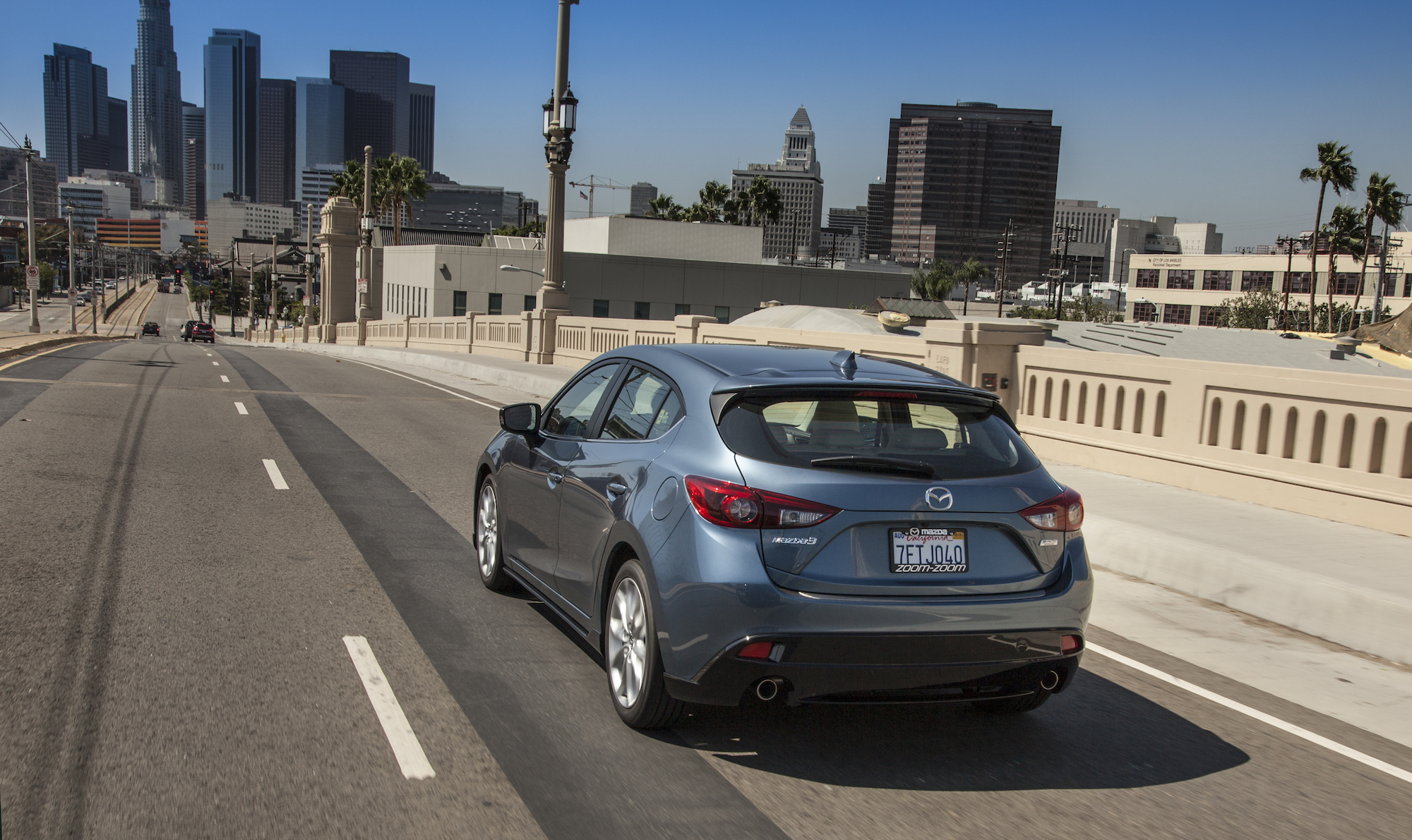 A grayish blue metallic 2016 Mazda3 five-door hatchback traveling over a bridge toward a city