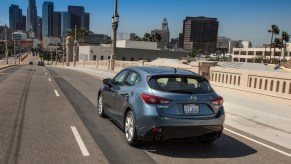 A grayish blue metallic 2016 Mazda3 five-door hatchback traveling over a bridge toward a city