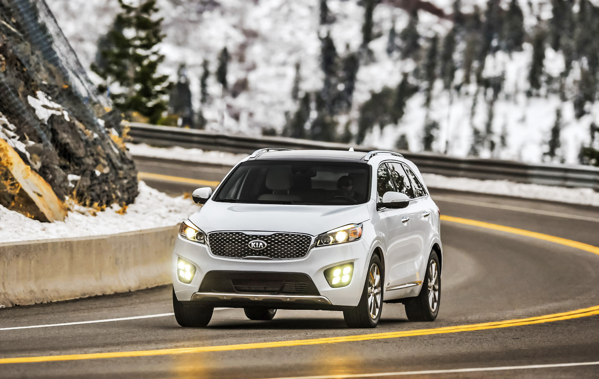 A white 2017 Kia Sorento SUV travels through a curve in a two-lane highway through snow-covered mountains
