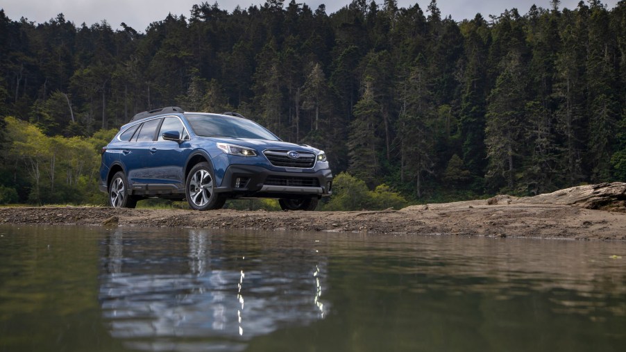 A blue 2020 Subaru Outback midsize SUV parked by a lake and pine trees