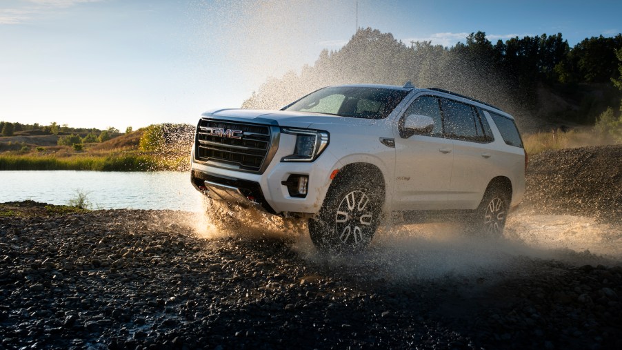A white 2021 GMC Yukon AT4 full-size SUV travels on wet gravel beside a lake and rolling green hills