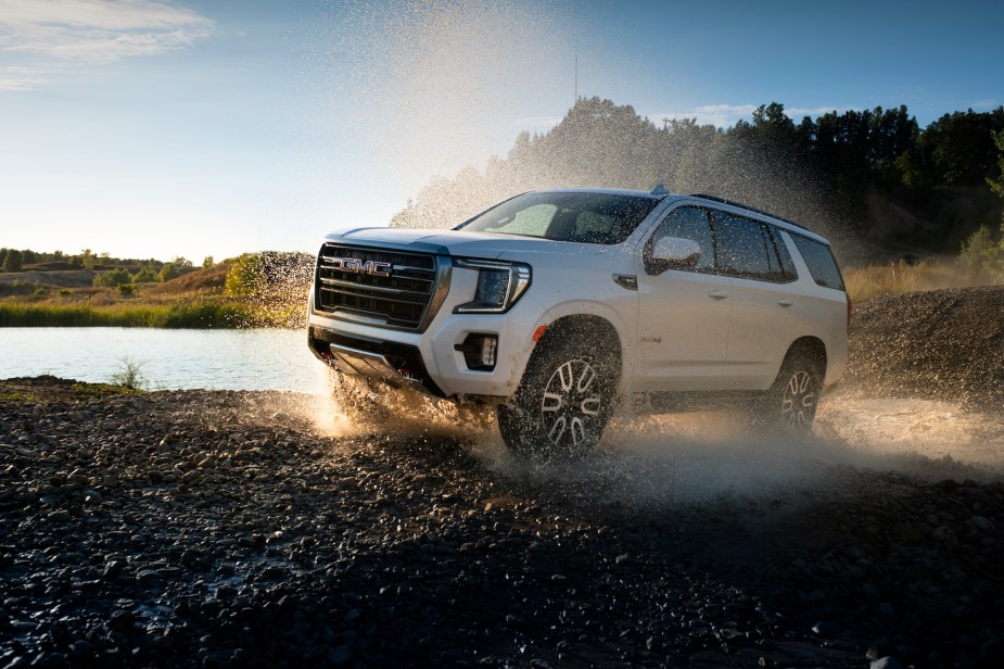 A white 2021 GMC Yukon AT4 full-size SUV travels on wet gravel beside a lake and rolling green hills