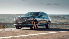A dark-green 2021 Genesis GV80 luxury midsize SUV parked next to a guardrail overlooking forest-covered hills