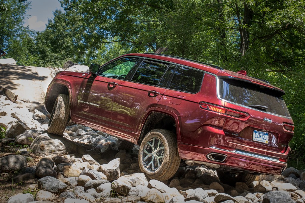 The rear 3/4 view of a red 2021 Jeep Grand Cherokee L Overland with Off-Road Group Package crawling up a rocky forest hill