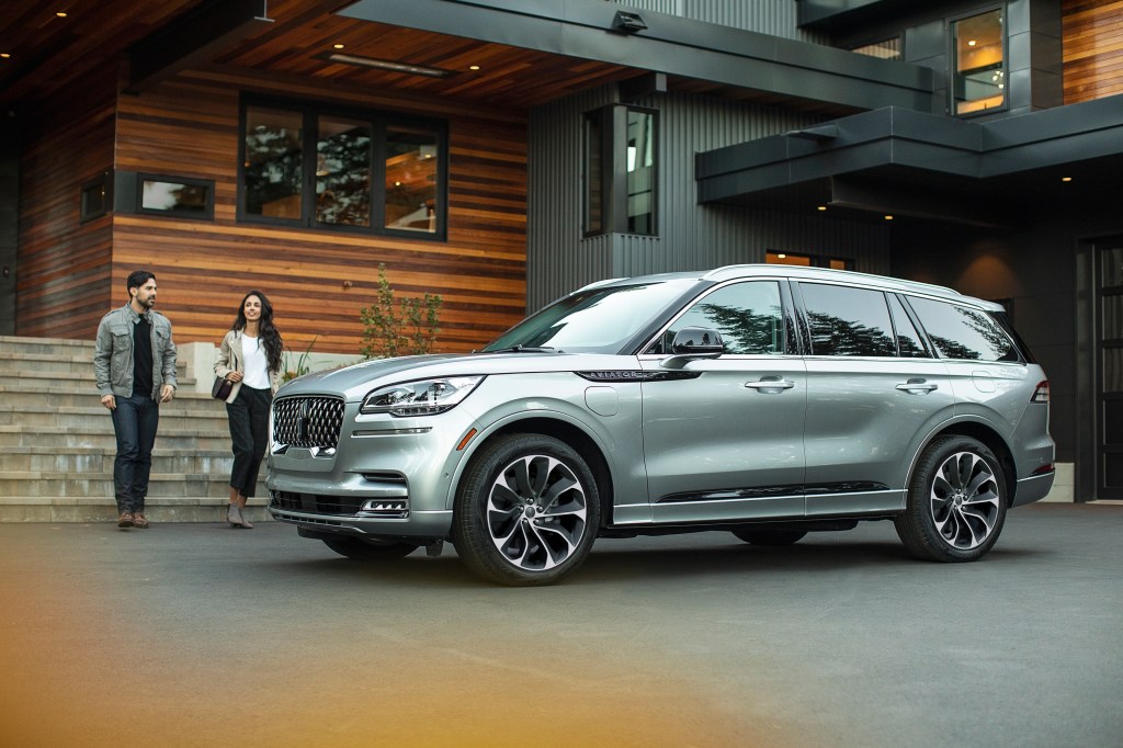 Two people walk up to a silver 2021 Lincoln Aviator Grand Touring plug-in hybrid parked by a wood-and-stone building
