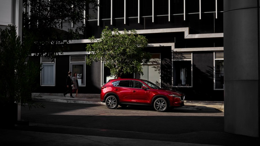 A red metallic 2021 Mazda CX-5 compact SUV parked on a city street as two office workers pass by on a sidewalk