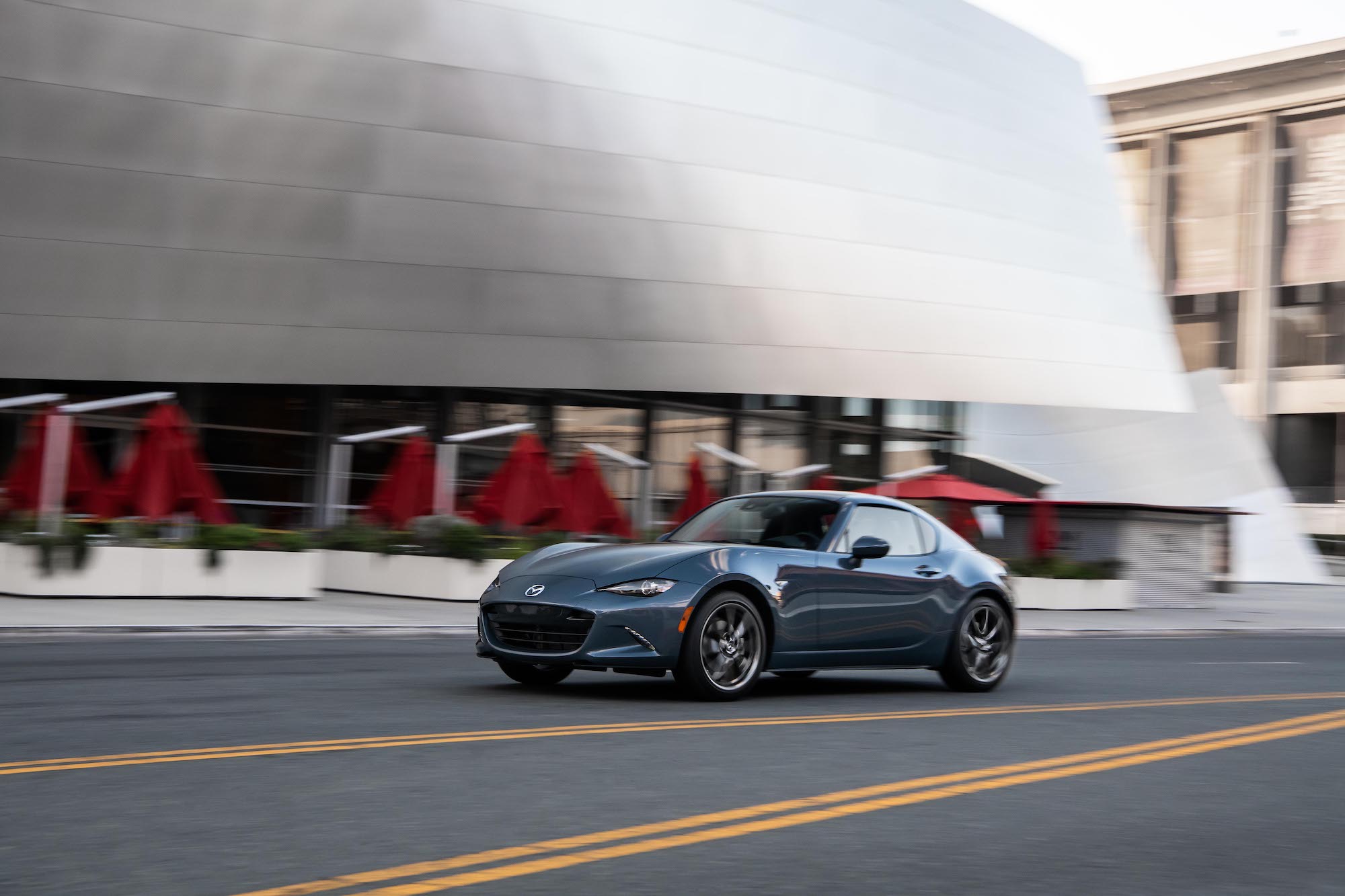 A 2021 Mazda MX-5 Miata RF sports car, with Polymetal Gray exterior paint, travels past a silver city building