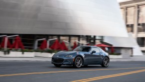A 2021 Mazda MX-5 Miata RF sports car, with Polymetal Gray exterior paint, travels past a silver city building