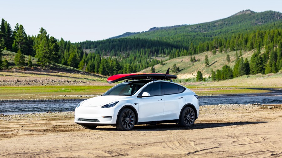 A white 2021 Tesla Model Y parked on the sand next to a river with pine trees and mountains in the distance