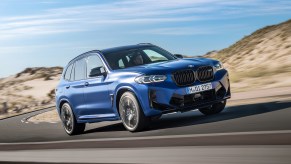A blue 2022 BMW X3 M Competition luxury compact SUV traveling on a paved road through arid hills