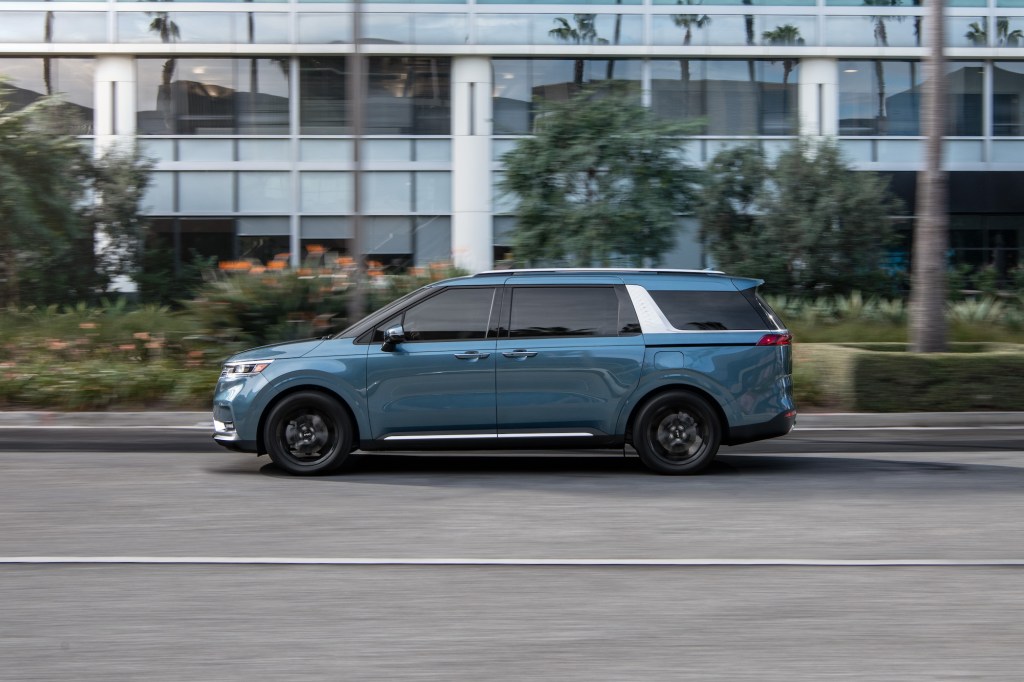 A blue 2022 Kia Carnival, which resembles an SUV and a minivan, travels on a street past palm trees and a modern building
