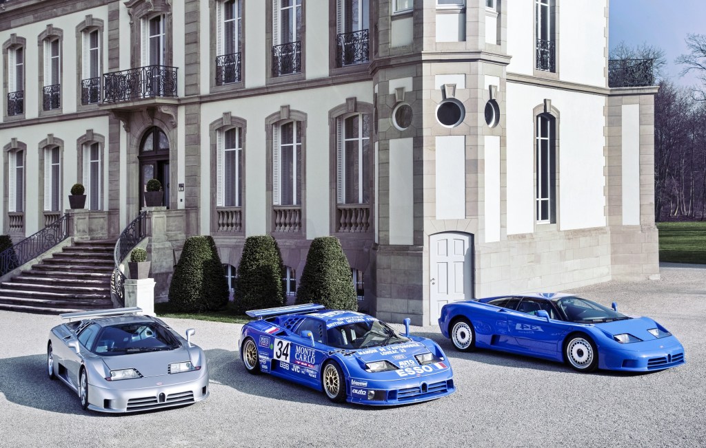 A silver Bugatti EB110 SS next to a blue EB110 race car next to a blue EB110 GT by the Molsheim, France chateau