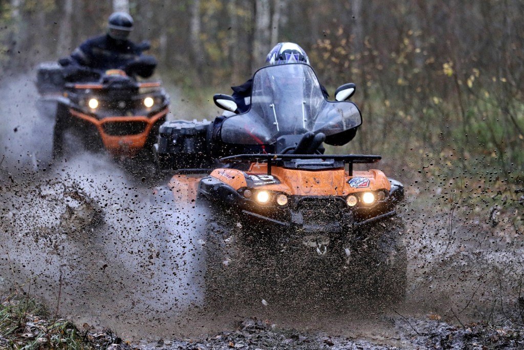 Two people in helmets ride orange ATVs through mud in a wooded area