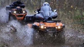 Two people in helmets ride orange ATVs through mud in a wooded area