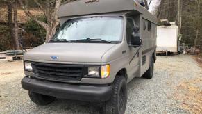 Ford E350 minibus camper conversion in a grayish green, parked on a gravel drive.