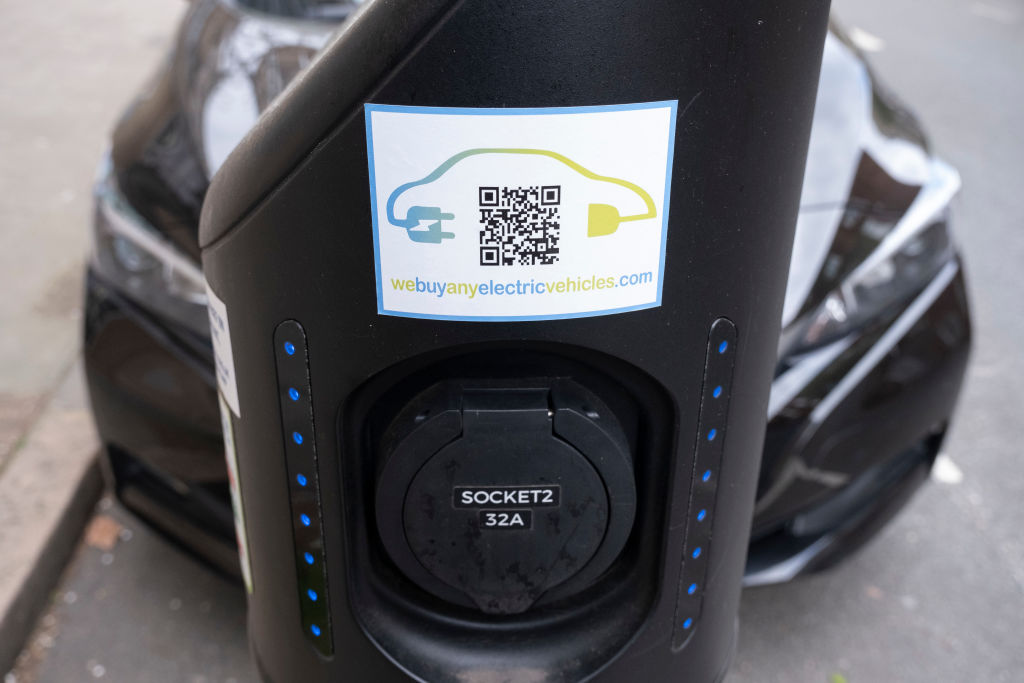 An EV charger in the foreground with a blurry EV in the background on a street