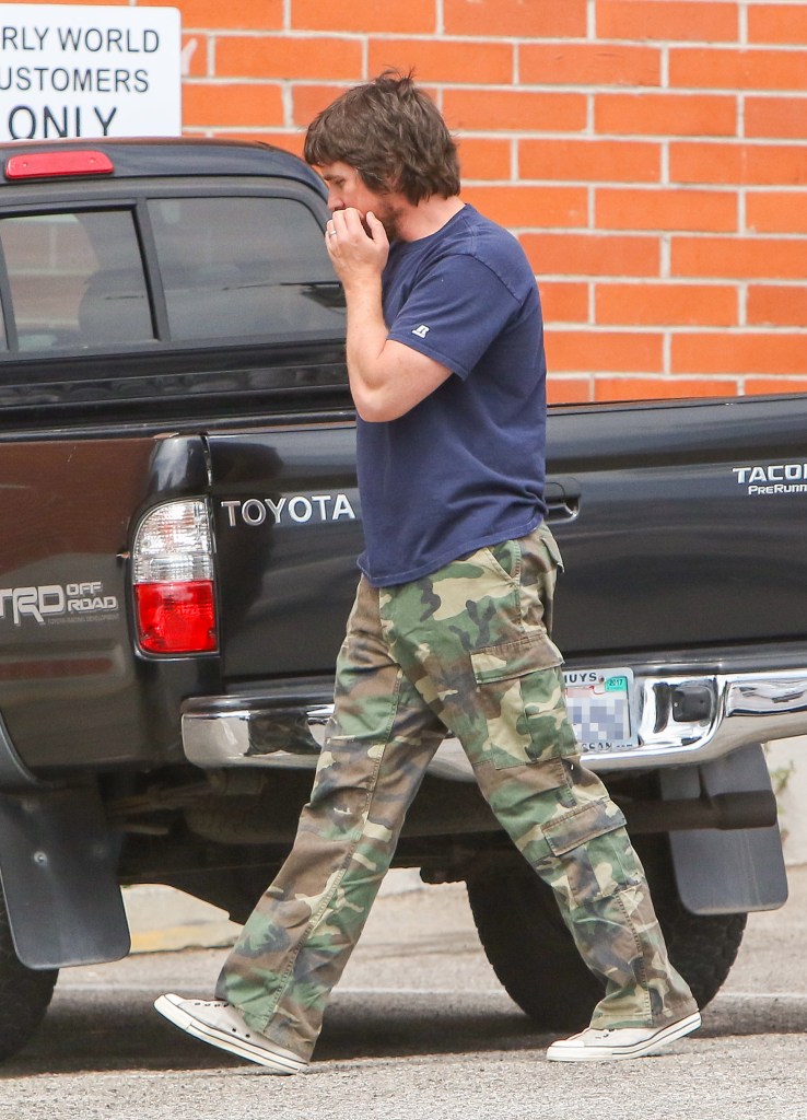 Actor Christian Bale walks behind his black 2003 Toyota Tacoma TRD Off-Road PreRunner V6 in camo cargo pants and white converse sneakers