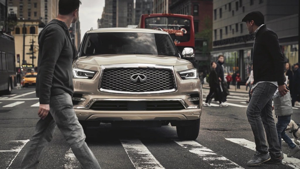 A beige 2021 Infiniti QX80 stopped at a crosswalk as pedestrians walk in front of it. 