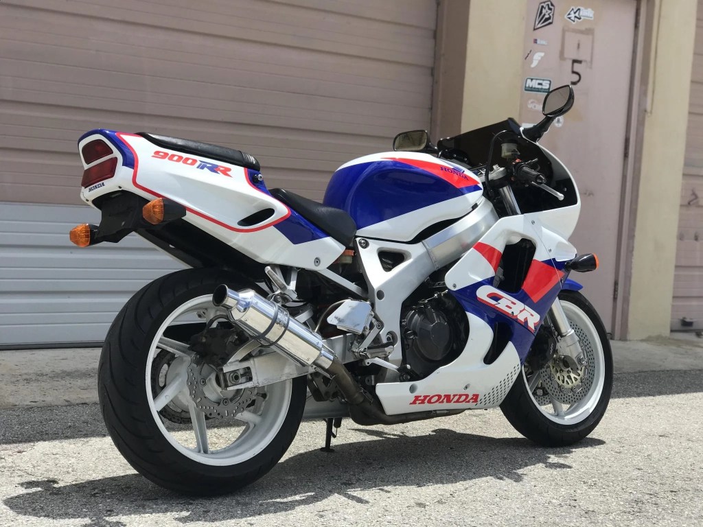 The rear 3/4 view of a modified white-red-and-blue 1993 Honda CBR900RR Fireblade by a tan garage