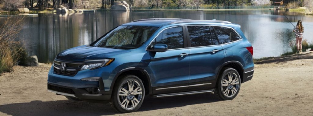 A blue 2021 Honda Pilot parked at a lake while a girl fishes in the background.