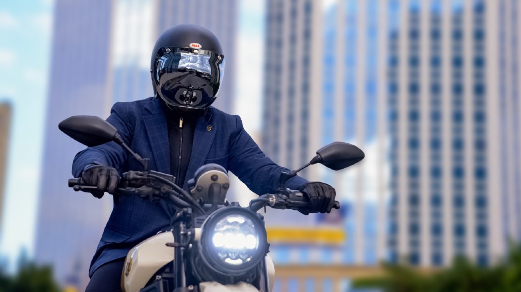 A black-helmeted rider on a white motorcycle commuting in a checkered-blue Brummell Blazer through a city