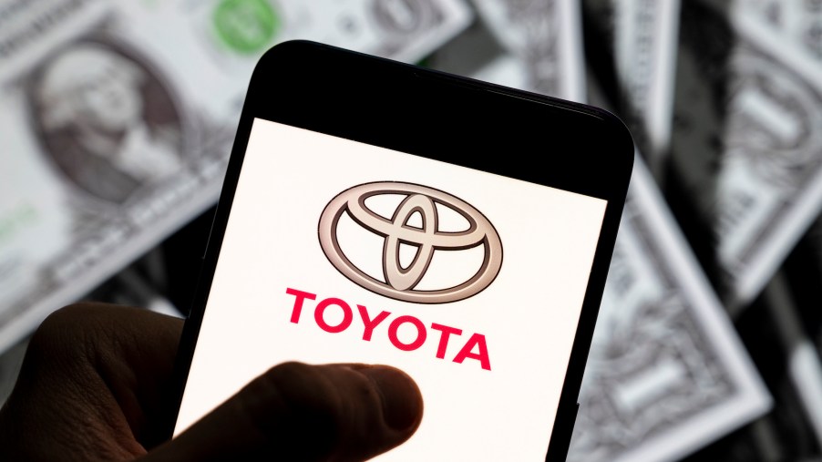 A photo illustration of the Toyota logo displayed on a smartphone screen with $1 bills in the background