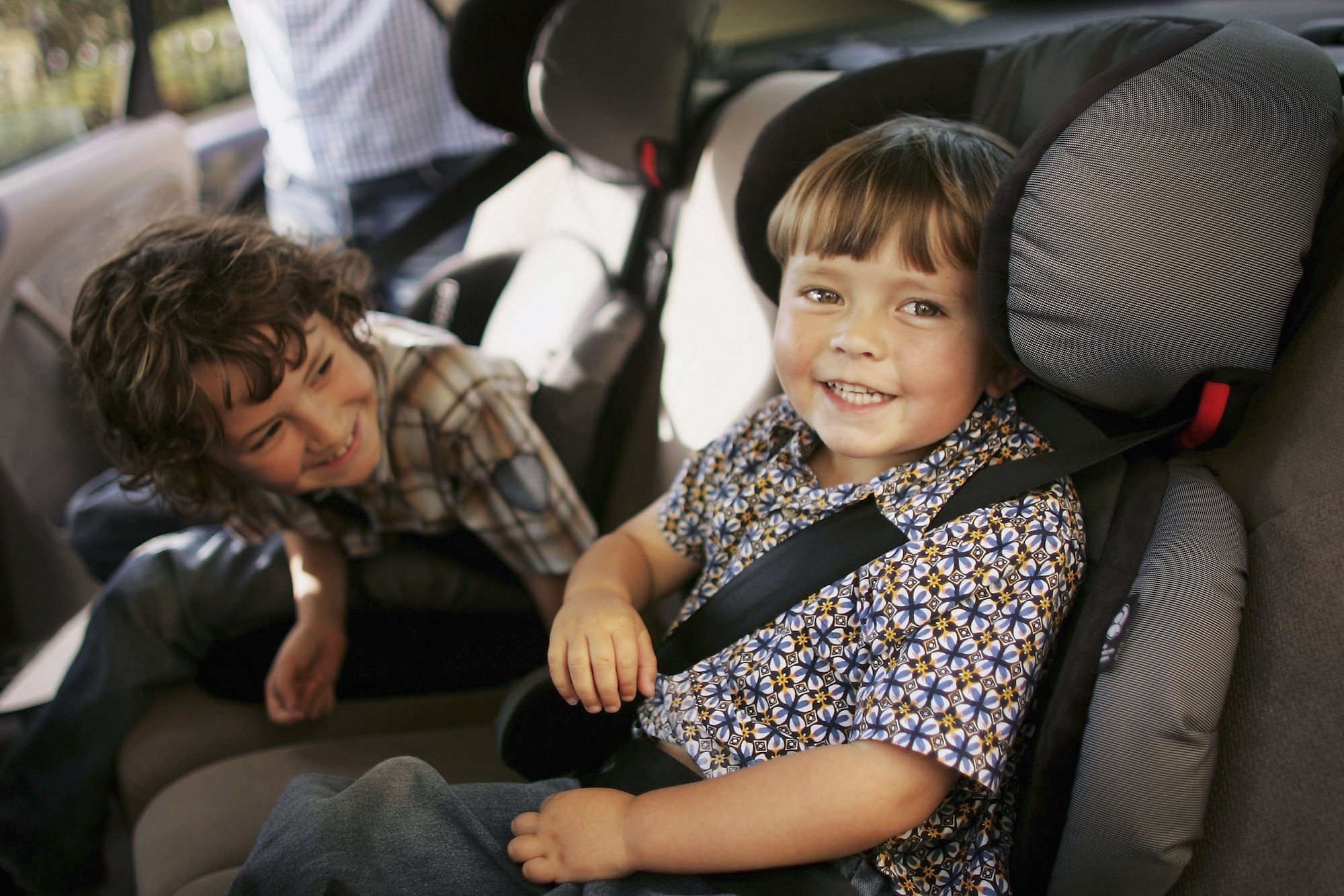 Two young children sit in car seats in the backseat of a vehicle