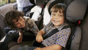 Two young children sit in car seats in the backseat of a vehicle