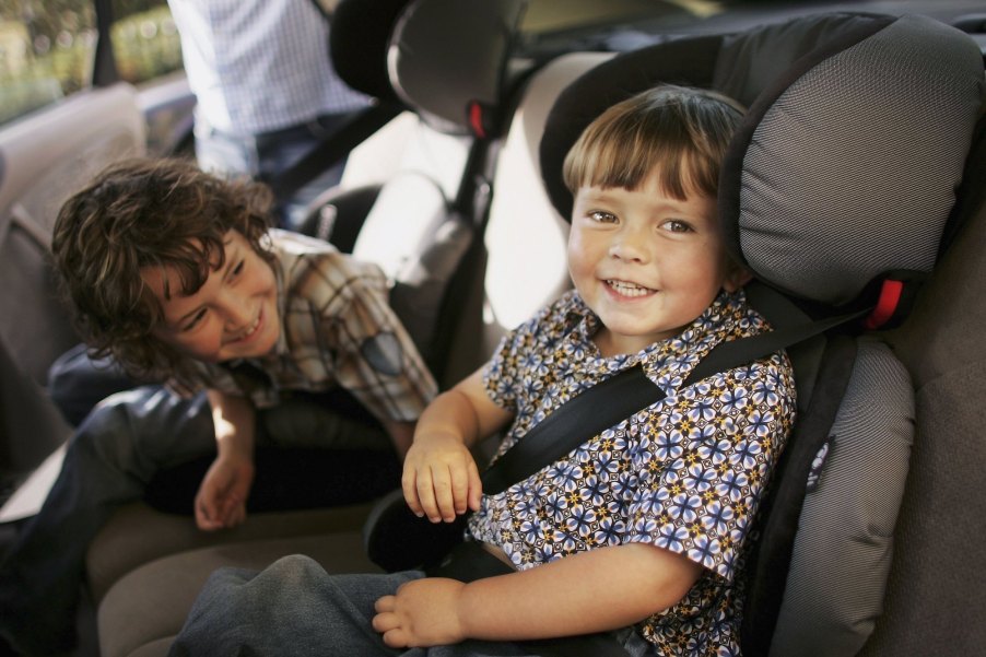 Two young children sit in car seats in the backseat of a vehicle